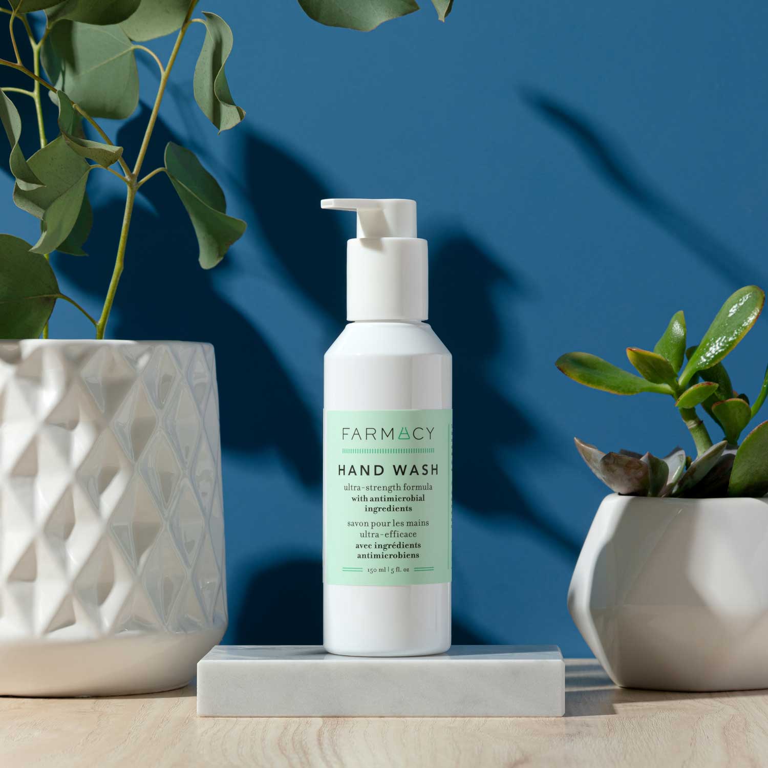 A bottle of Farmacy's Hand Wash sitting in the middle of two green potted plants